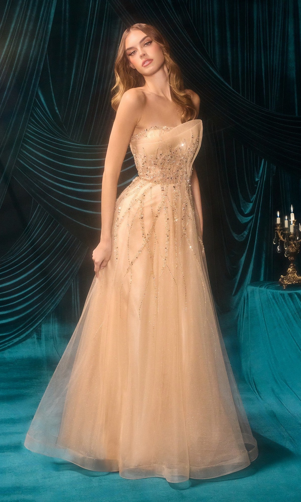 A woman poses in the Andrea and Leo A1491, a floor-length nude tulle prom dress with sparkling bead embellishments, standing before teal velvet drapes and a candelabra.